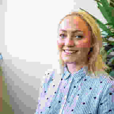 Art director headshot, medium blonde hair with blue shirt, white background, next to plant.